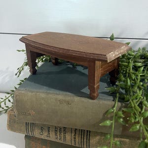 May include: A miniature brown wooden table with a rectangular top and ornate legs. The table is set on top of a stack of old books. The table is approximately 6 inches long and 3 inches wide.