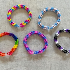 May include: Five colourful loom band bracelets. The bracelets are made of small, woven rubber bands in various colour combinations, including rainbow, blue and white, purple and blue, and black and white. The bracelets are shown on a neutral background.
