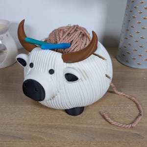 May include: White ceramic yarn bowl shaped like a yak with brown horns. The bowl is filled with a ball of light brown yarn and two knitting needles.
