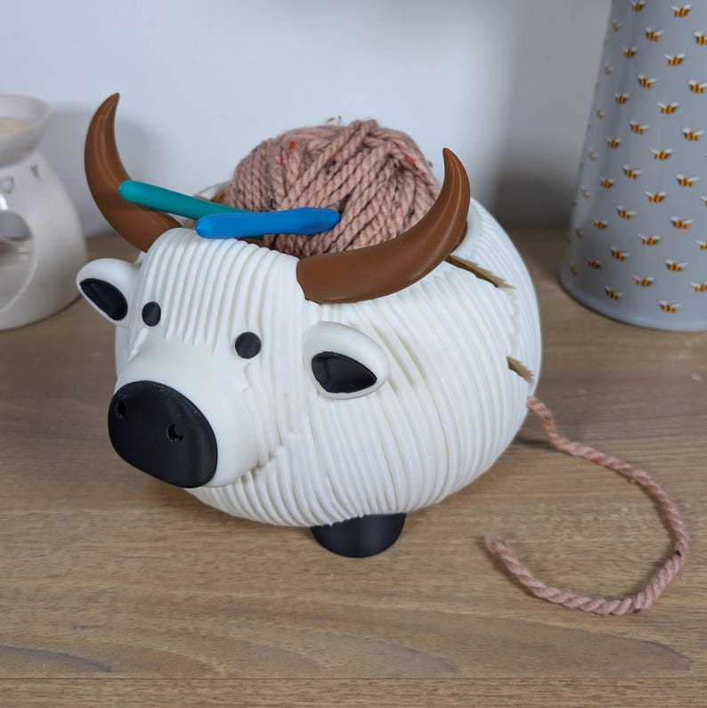 May include: White ceramic yarn bowl shaped like a yak with brown horns. The bowl is filled with a ball of light brown yarn and two knitting needles.
