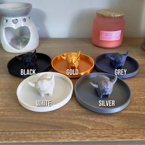 May include: A collection of Highland cow figurines in various colors: black, gold, grey, white, and silver. Each cow sits on a matching round tray. The colors are labeled with text. A candle and oil diffuser are in the background.