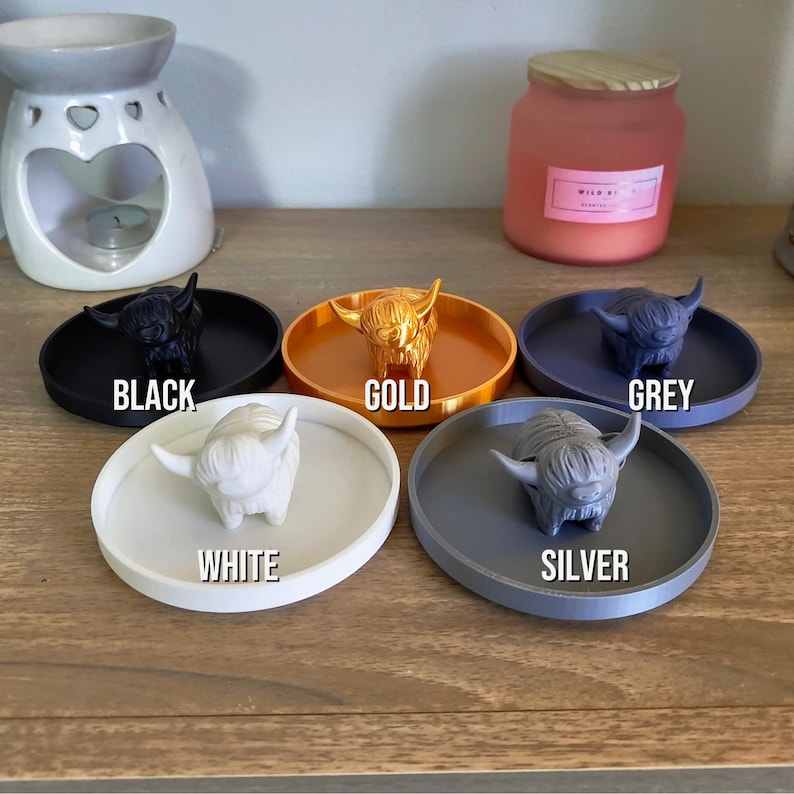 May include: A collection of Highland cow figurines in various colors: black, gold, grey, white, and silver. Each cow sits on a matching round tray. The colors are labeled with text. A candle and oil diffuser are in the background.