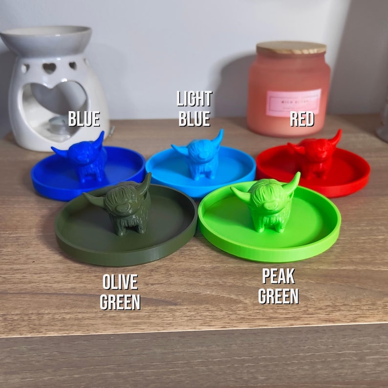 May include: A collection of colorful Highland cow-shaped trinket dishes in blue, light blue, red, olive green, and peak green. Each dish features a small cow figurine in the center. The word "BLUE", "LIGHT BLUE", "RED", "OLIVE GREEN", and "PEAK GREEN" are written on the image.