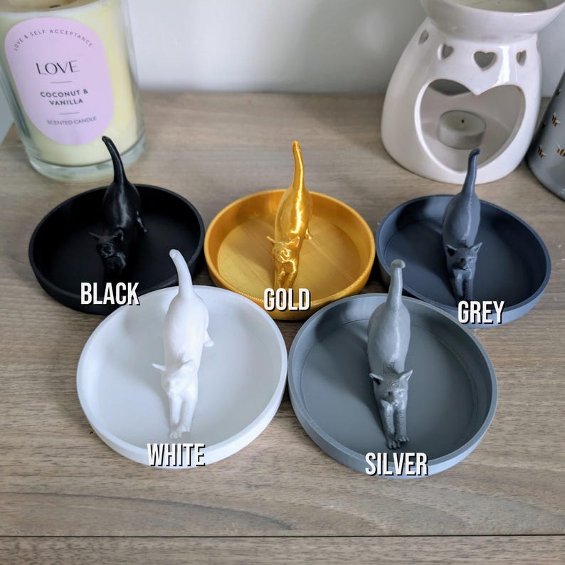 May include: Five ring dishes with a cat figurine in the center. The dishes are in black, gold, white, silver, and grey.