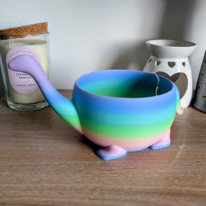 May include: A rainbow-colored ceramic bowl shaped like a dinosaur with a long neck and legs. The bowl is on a wooden surface.