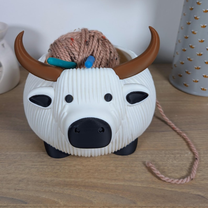 May include: White ceramic yarn bowl shaped like a cow with brown horns. The bowl is filled with a ball of brown yarn and two knitting needles.