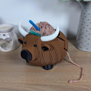 May include: A brown yarn bowl shaped like a Highland cow with white horns. The bowl has a pink ball of yarn inside and two knitting needles sticking out of the top.