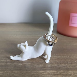 May include: White ceramic cat ring holder with three rings on its tail. The cat is in a stretched out pose with its head turned to the side.