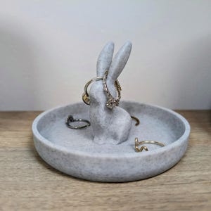 May include: A grey ceramic ring holder shaped like a rabbit with a speckled finish. The rabbit is holding several rings on its ears and there are more rings in the dish.