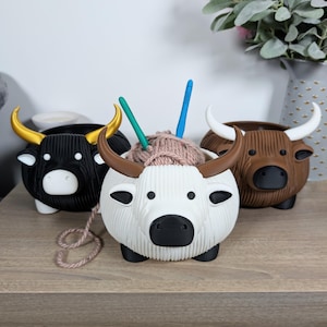 May include: Three ceramic yarn bowl holders shaped like bulls. The bowls are white, black, and brown. The white bowl has yarn and knitting needles inside.