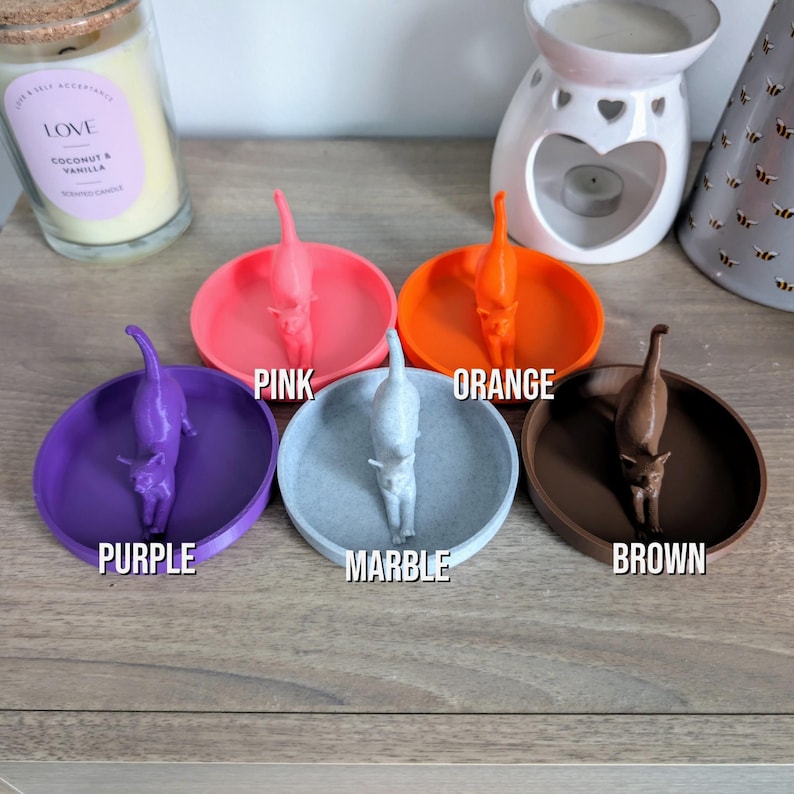 May include: Five ring dishes with a cat figurine on each. The dishes are in the colors pink, orange, purple, marble, and brown. The dishes are labeled with their color.