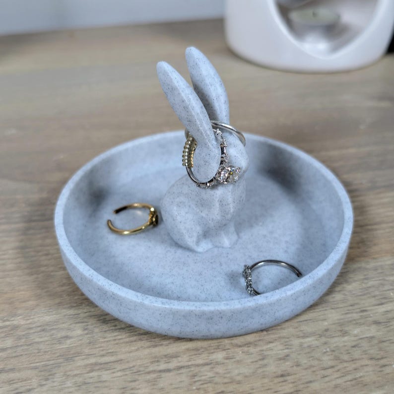 May include: A grey ceramic ring holder shaped like a rabbit with a speckled finish. The rabbit is holding rings on its ears and there are additional rings in the dish.