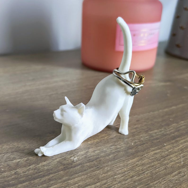 May include: White ceramic cat ring holder with a ring on its tail. The cat is in a stretched out pose, with its head turned to the side. The ring holder is on a wooden surface.