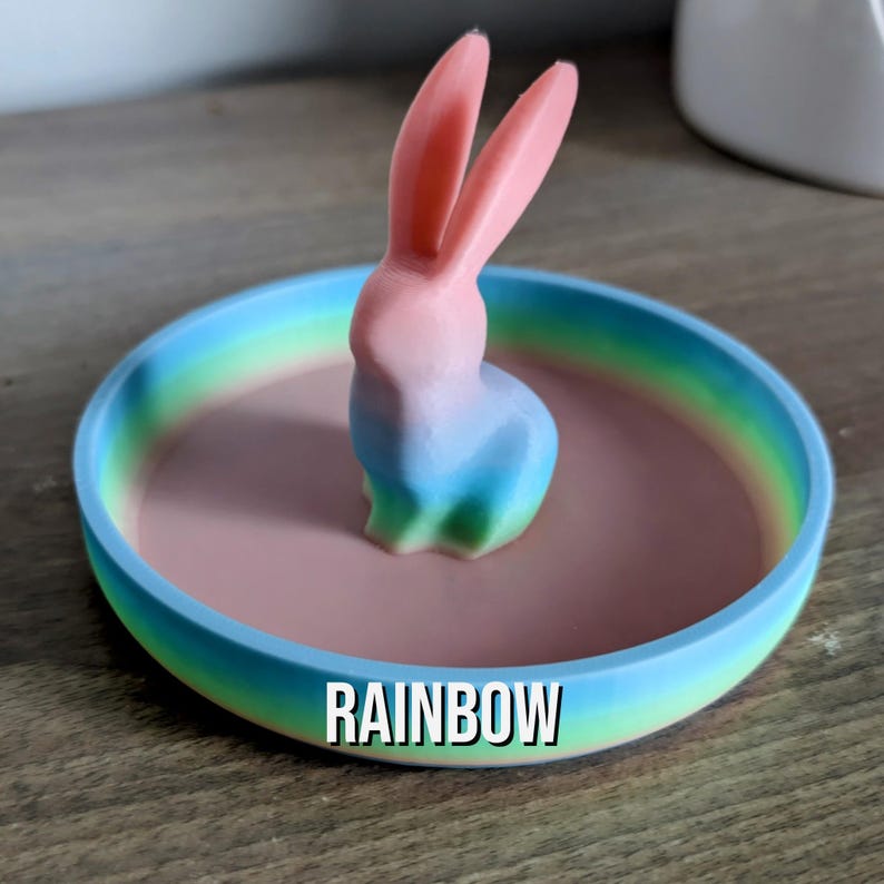 May include: A rainbow coloured ring dish with a pastel pink interior and a 3D printed ombre bunny figurine in the centre. The dish has the word "RAINBOW" printed on the bottom.