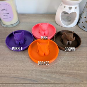 May include: Four small ceramic dishes with elephant figurines in the center. The dishes are in the colors purple, pink, orange, and brown.