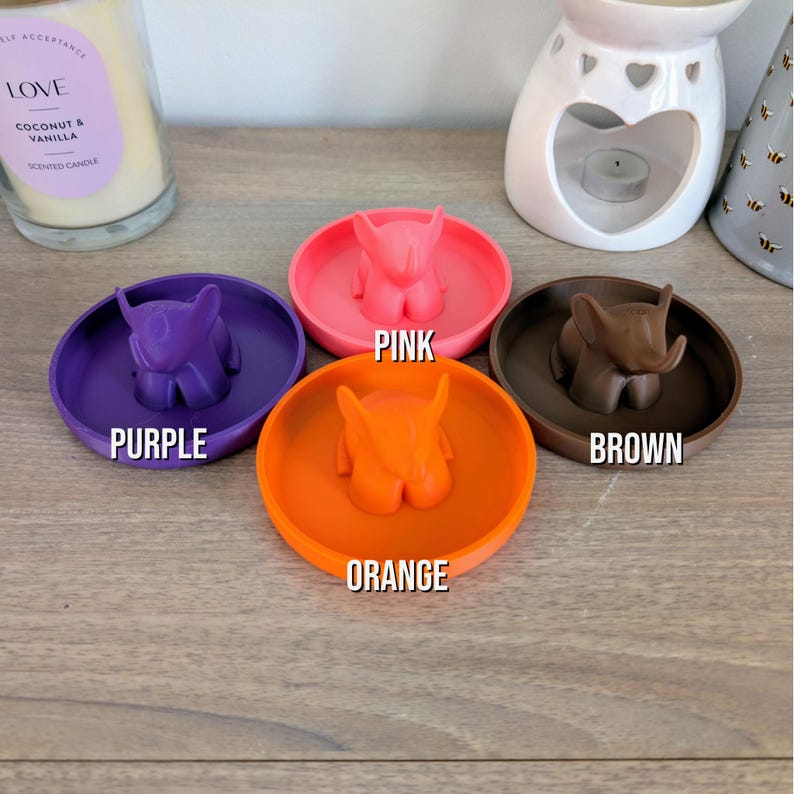May include: Four small ceramic dishes with elephant figurines in the center. The dishes are in the colors purple, pink, orange, and brown.