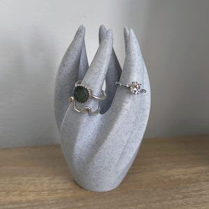 Op de afbeelding: Een witte keramische ringenhouder in de vorm van een hand met drie vingers. De houder heeft een gespikkelde textuur en houdt drie ringen vast, een met een groene steen en twee zilveren banden.