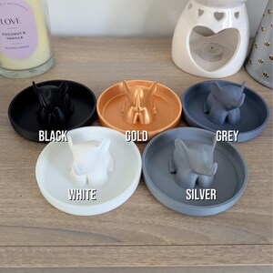 May include: Five small ceramic dishes with elephant figurines in different colors: black, gold, gray, white, and silver. The dishes are on a wooden surface.