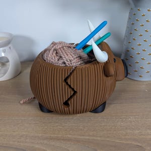 May include: Brown ceramic yarn bowl shaped like a bull with a crack down the side. The bowl is filled with pink yarn and has three knitting needles sticking out of the top.