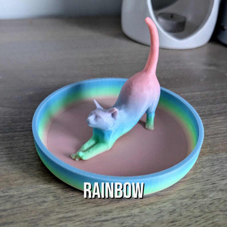 May include: A rainbow colored cat figurine stretches out on a ring dish with a rainbow gradient. The dish is made of a smooth, light blue material. The text "RAINBOW" is printed below the dish.