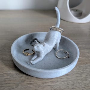 May include: A gray ceramic cat ring holder with a white speckled finish. The cat is in a playful pose with its tail raised. The holder is filled with several rings.