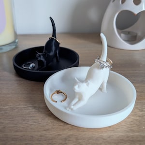 May include: Two cat-shaped ring holders, one black and one white, are displayed on their respective dishes. The black cat is sitting up with its tail curled around its body, while the white cat is lying down with its tail curled around its body. Both cats have rings on their tails.