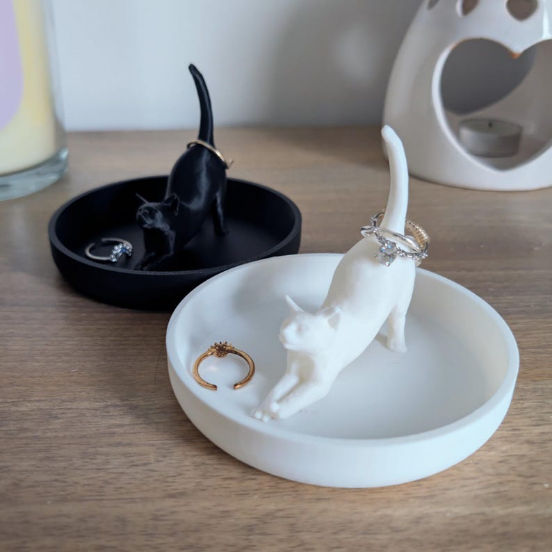May include: Two cat-shaped ring holders, one black and one white, are displayed on their respective dishes. The black cat is sitting up with its tail curled around its body, while the white cat is lying down with its tail curled around its body. Both cats have rings on their tails.