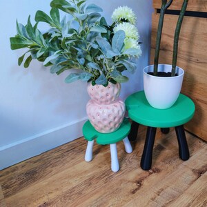 May include: A pink strawberry-shaped vase with green leaves and white flowers sits on a small green stool with white legs. Another green stool with black legs holds a white pot with a plant.