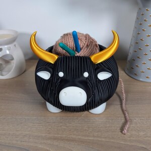 May include: Black yarn bowl shaped like a bull with gold horns. The bowl is filled with a ball of yarn and knitting needles.
