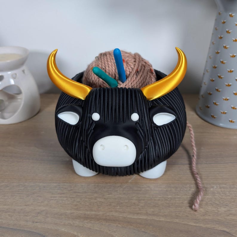 May include: Black yarn bowl shaped like a bull with gold horns. The bowl is filled with a ball of yarn and knitting needles.