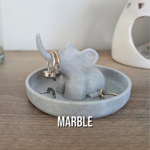 May include: A gray marble elephant ring holder with a small dish. The elephant has a raised trunk and is holding rings on its trunk and ears. The dish is round and has a textured surface. The word "MARBLE" is printed on the dish.