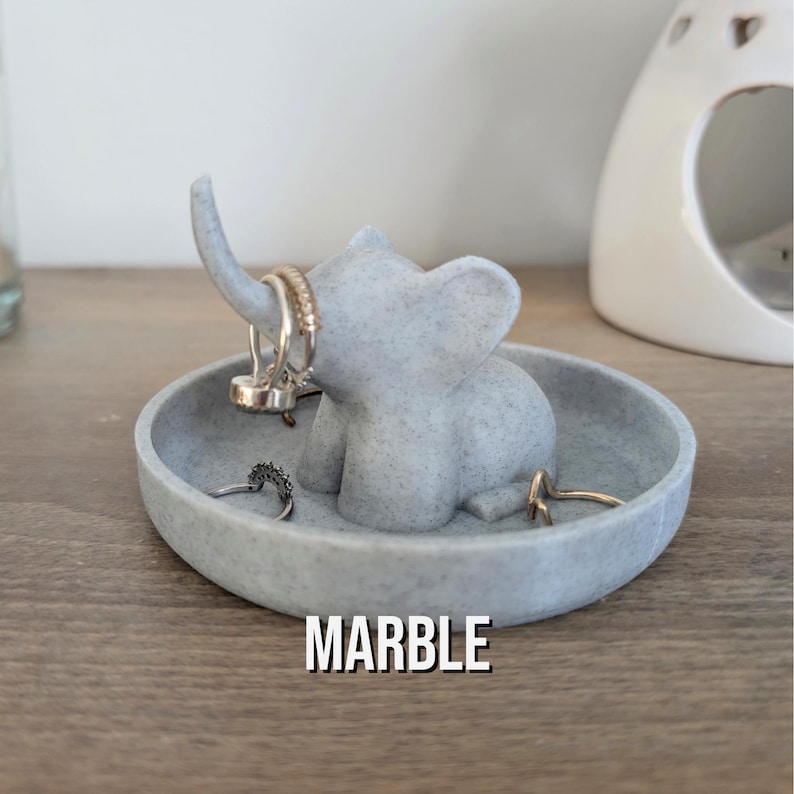 May include: A gray marble elephant ring holder with a small dish. The elephant has a raised trunk and is holding rings on its trunk and ears. The dish is round and has a textured surface. The word "MARBLE" is printed on the dish.