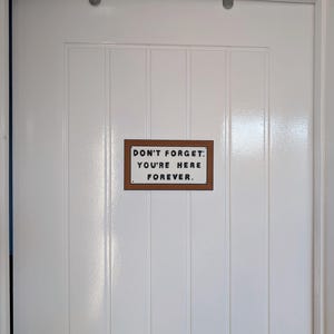 May include: A white door with a brown sign that reads "Don't forget: You're here forever."