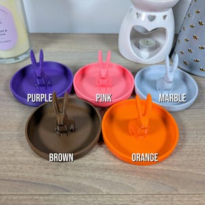 May include: Five ring dishes with a bunny rabbit figurine on top. The dishes are in the colours purple, pink, brown, orange, and light grey. The dishes are labelled with the colour of each dish.