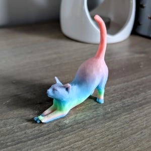 May include: A rainbow-colored cat figurine stretching with its tail raised in the air. The figurine is made of a smooth, glossy material and has a vibrant color gradient from pink to blue.