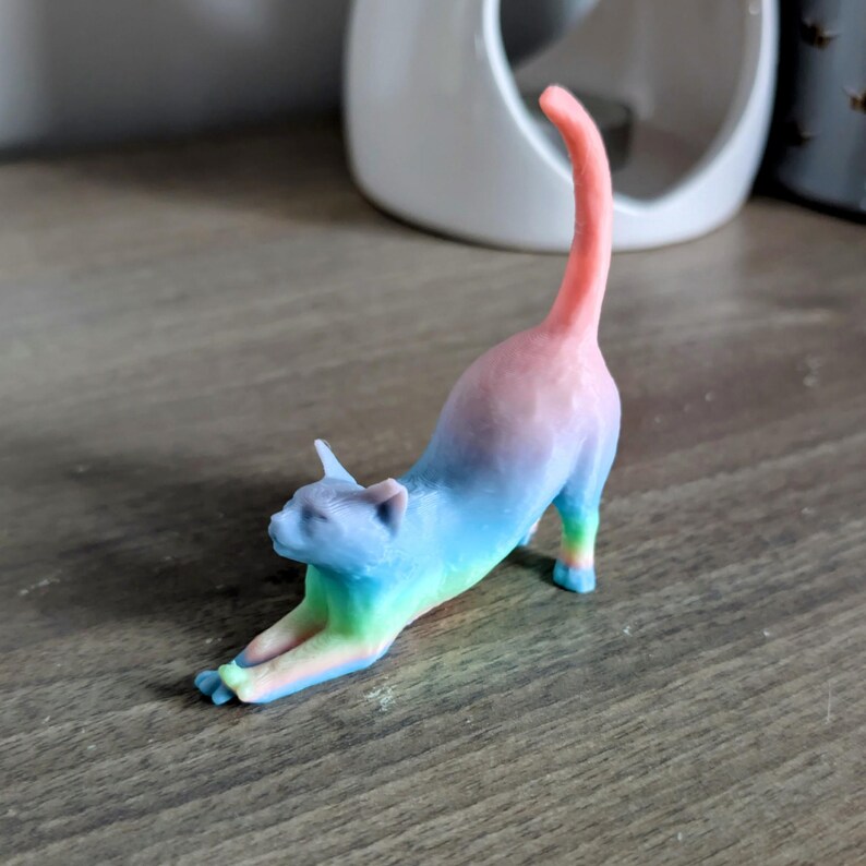 May include: A rainbow-colored cat figurine stretching with its tail raised in the air. The figurine is made of a smooth, glossy material and has a vibrant color gradient from pink to blue.