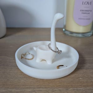 May include: White ceramic ring holder shaped like a dinosaur with a long neck. The dinosaur is holding a ring on its neck. There are other rings in the dish.