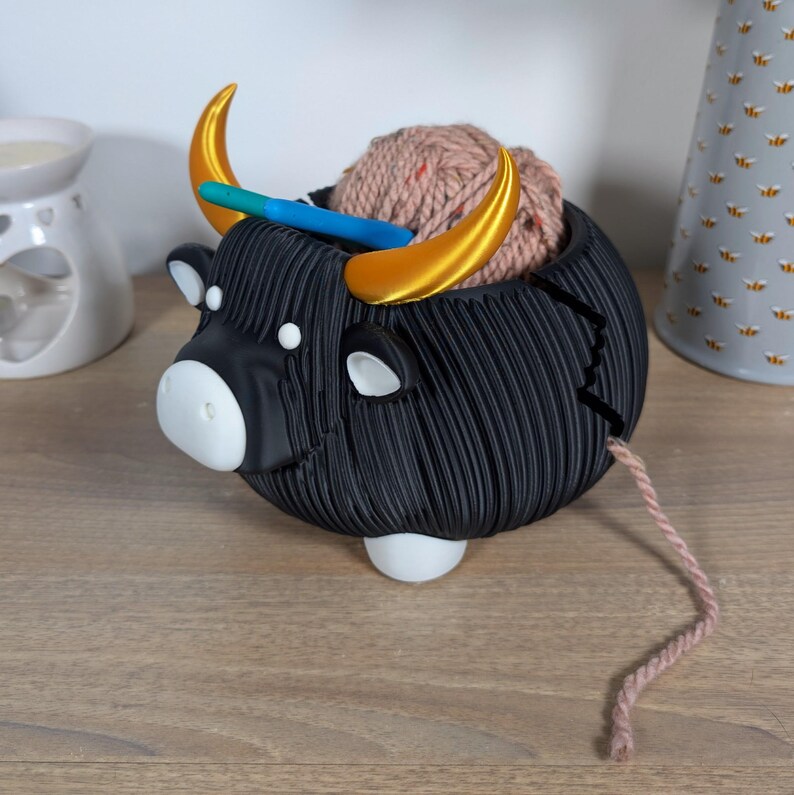May include: Black yarn bowl shaped like a Highland cow with gold horns. The bowl is holding a ball of pink yarn and a knitting needle.