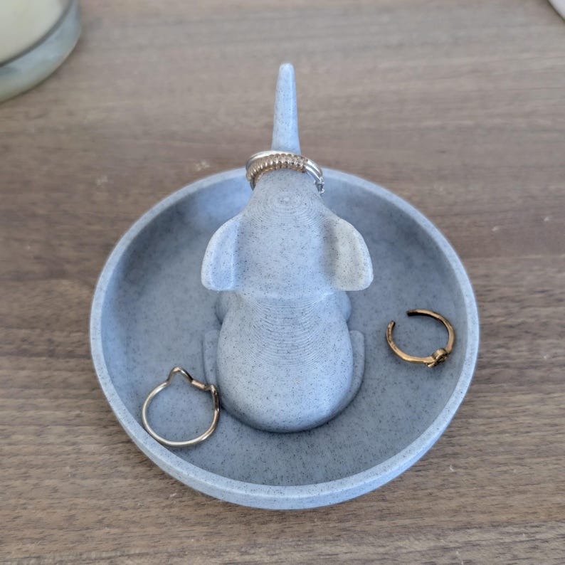 May include: A gray ceramic elephant ring holder with a small dish. The elephant is sitting up with its trunk raised and has a ring on its trunk. There are two other rings in the dish.