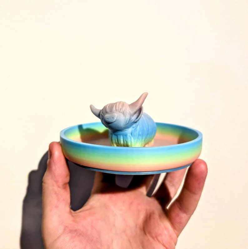 May include: A small, colorful dish with a Highland cow figurine. The dish has a rainbow gradient of colors, and the cow is blue and gray. The figurine is a decorative item.