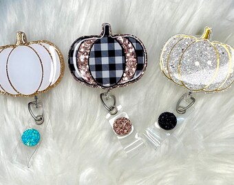 May include: Three pumpkin-shaped badge reels with glitter accents. One is white with gold trim, another has a black and white plaid pattern with rose gold glitter, and the third is silver glitter with gold trim. Each reel has a different colored glitter ball.