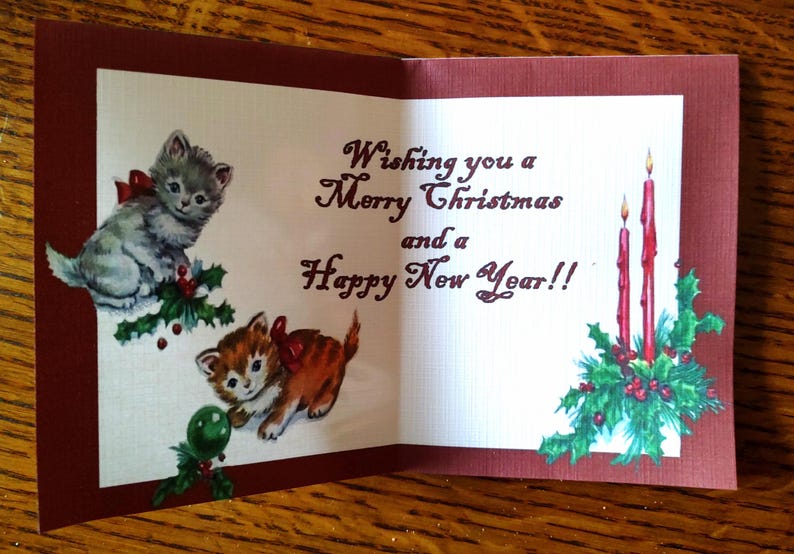 K&ouml;nnte beinhalten: Eine Vintage-Weihnachtskarte mit Illustrationen von zwei K&auml;tzchen mit roten Schleifen, Stechpalmen und brennenden Kerzen. Die Karte tr&auml;gt die Aufschrift "Wishing you a Merry Christmas and a Happy New Year!!" Die Karte hat einen burgunderfarbenen Rand.