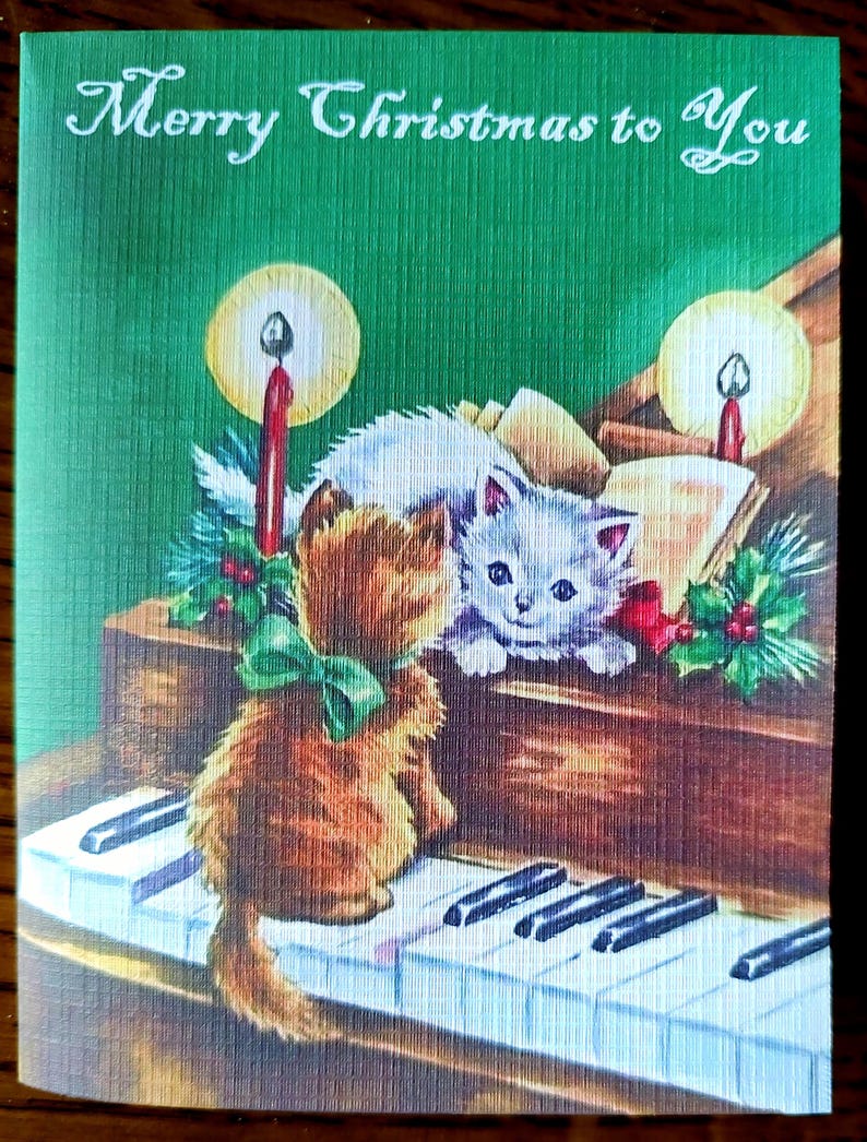 K&ouml;nnte beinhalten: Vintage Weihnachtskarte mit gr&uuml;n strukturiertem Hintergrund und dem Text "Merry Christmas to You". Die Karte zeigt Illustrationen von zwei K&auml;tzchen, Kerzen und einem Klavier. Ein K&auml;tzchen sitzt auf den Klaviertasten, das andere ruht auf dem Klavier.