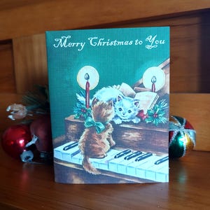 K&ouml;nnte beinhalten: Weihnachtskarte mit gr&uuml;nem Hintergrund und dem Text "Merry Christmas to You". Die Karte zeigt eine Vintage-Illustration von zwei K&auml;tzchen, die Klavier spielen, mit Kerzen und Stechpalmenakzenten.