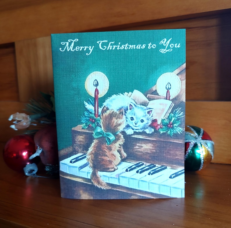 K&ouml;nnte beinhalten: Weihnachtskarte mit gr&uuml;nem Hintergrund und dem Text "Merry Christmas to You". Die Karte zeigt eine Vintage-Illustration von zwei K&auml;tzchen, die Klavier spielen, mit Kerzen und Stechpalmenakzenten.