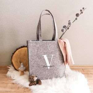 Könnte beinhalten: Graue Filztasche mit einem weißen Buchstaben V und dem Namen Vanessa darunter in Kursivschrift. Die Tasche liegt auf einem weißen, flauschigen Teppich mit einem kleinen Holzpilz und Tannenzapfen.