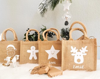 Può includere: Quattro piccole borse regalo in juta con disegni e nomi bianchi. Le borse sono decorate con Babbo Natale, un omino di pan di zenzero, una stella e una renna. Le borse sono appoggiate su una superficie bianca e soffice con due regali avvolti davanti a loro.