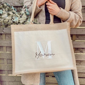 Könnte beinhalten: Eine beige Jute-Tragetasche mit einem weißen Canvas-Einsatz. Der Canvas-Einsatz zeigt einen großen weißen Buchstaben "M" und den Namen "Maren" in Kursivschrift unter dem Buchstaben.