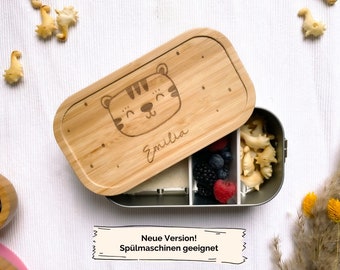 Peut inclure: Une boîte à lunch en bois avec un joli motif de tigre et le nom "Emilia" gravé sur le couvercle. La boîte à lunch est ouverte et montre un récipient en acier inoxydable avec des compartiments pour la nourriture. Le récipient contient des baies, des craquelins et une substance blanche à l'intérieur.