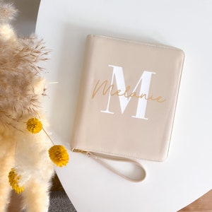 Könnte beinhalten: Ein beigefarbenes Ledernotizbuch mit einem weißen Buchstaben "M" und dem Namen "Melanie" in goldener Schrift.